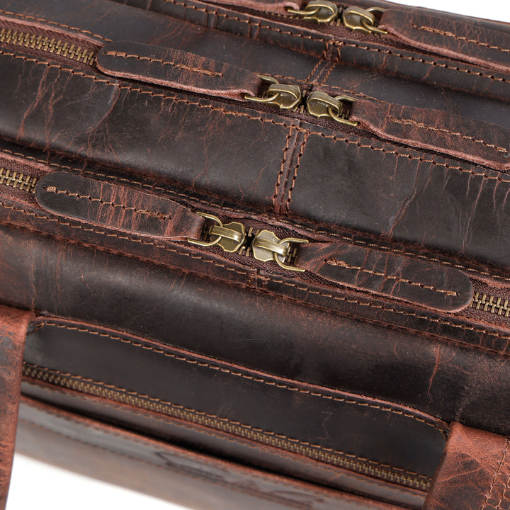 Close-up of a distressed brown leather bag showing stitched compartments, brass zippers, and decorative metal hardware.